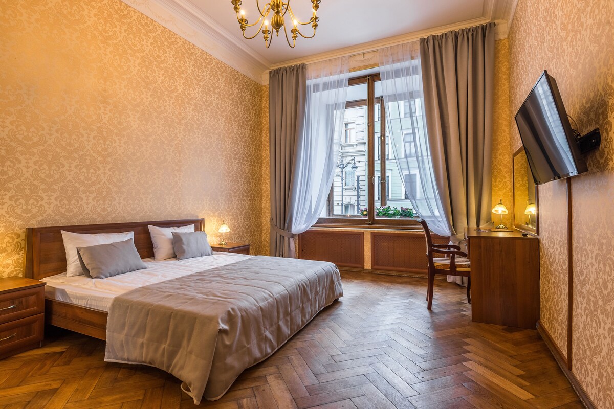 A hotel room with a large bed, wooden floor, a TV on the wall, and a window with curtains.
