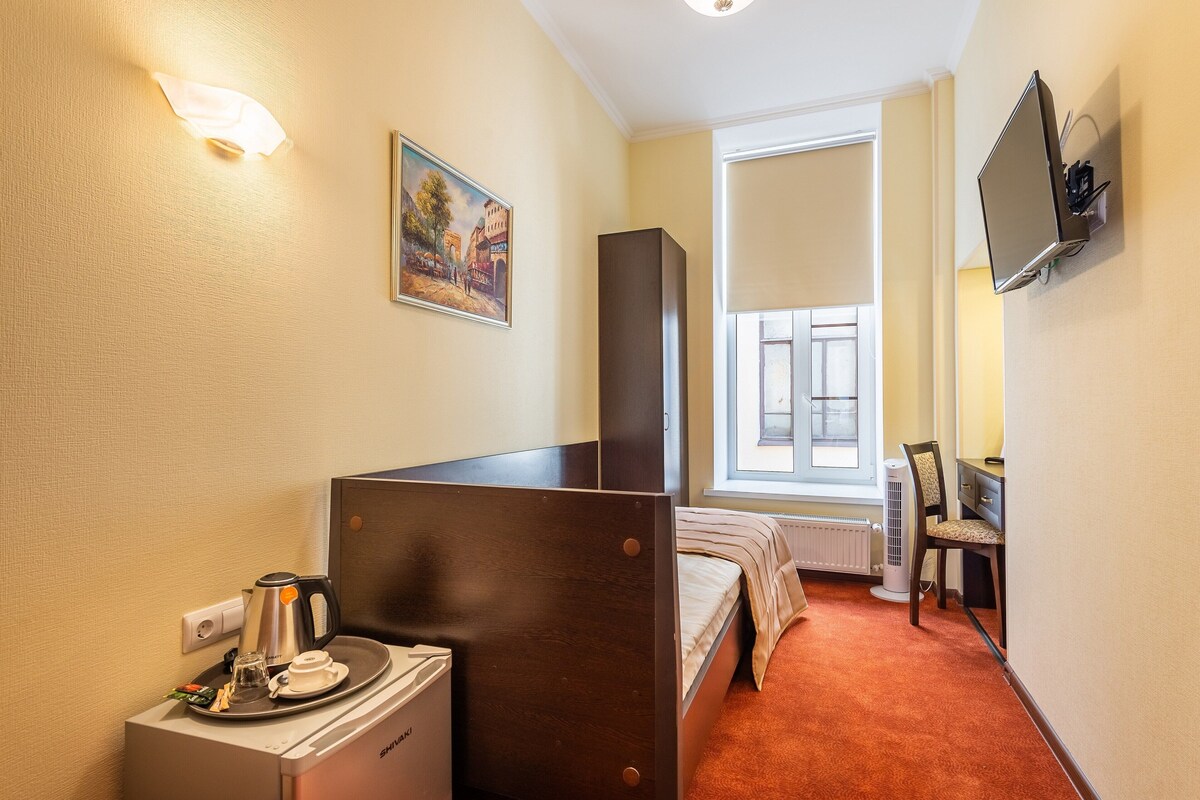 A hotel room with a single bed, a desk with a chair, a TV mounted on the wall, and a window with blinds.