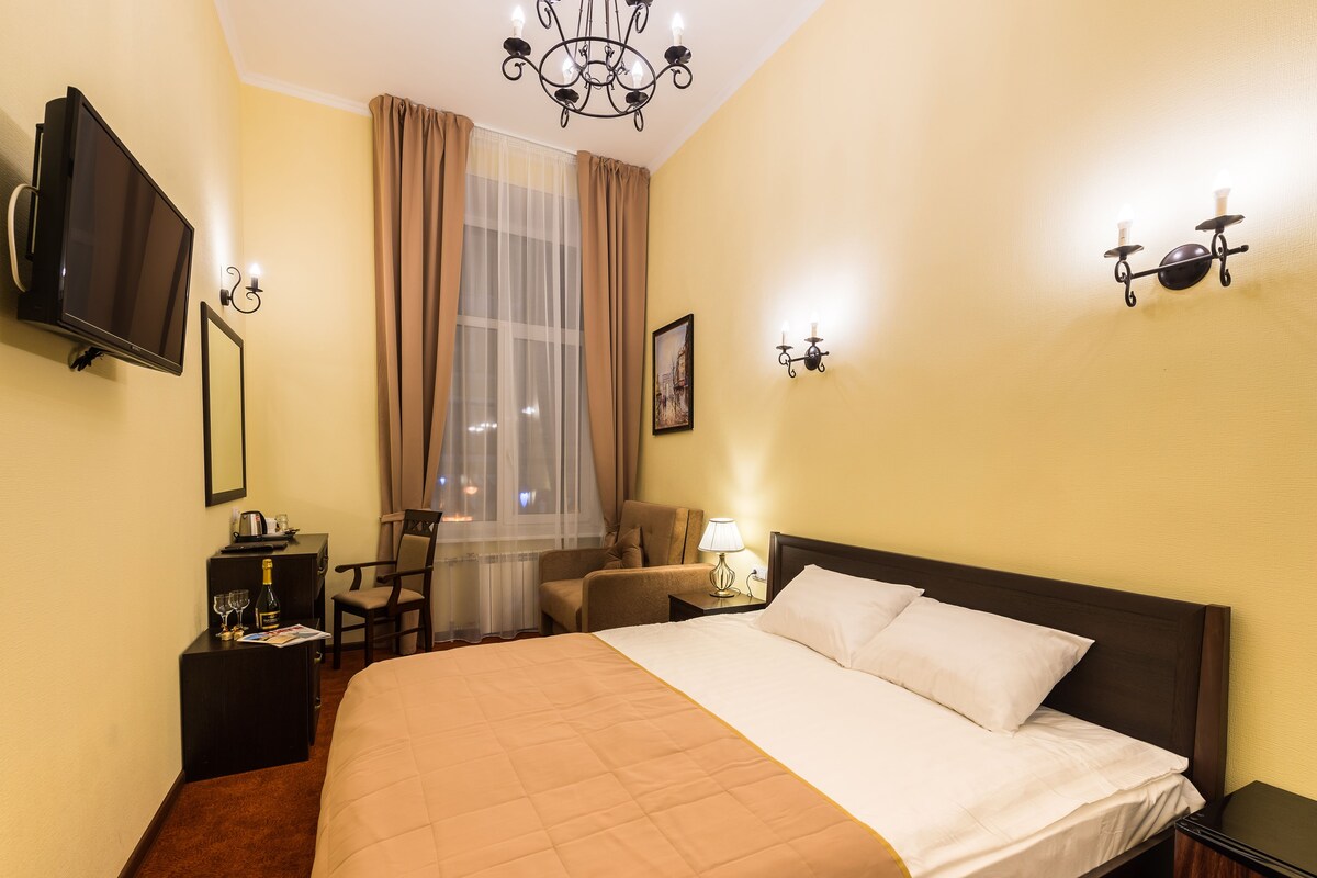 A hotel room with a bed, a desk with a television, a chair, a lamp, and a window with curtains.