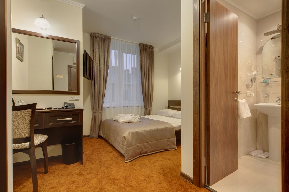 A hotel room with a bed, a desk with a chair, a bathroom with a sink and mirror, and a door leading to another room.