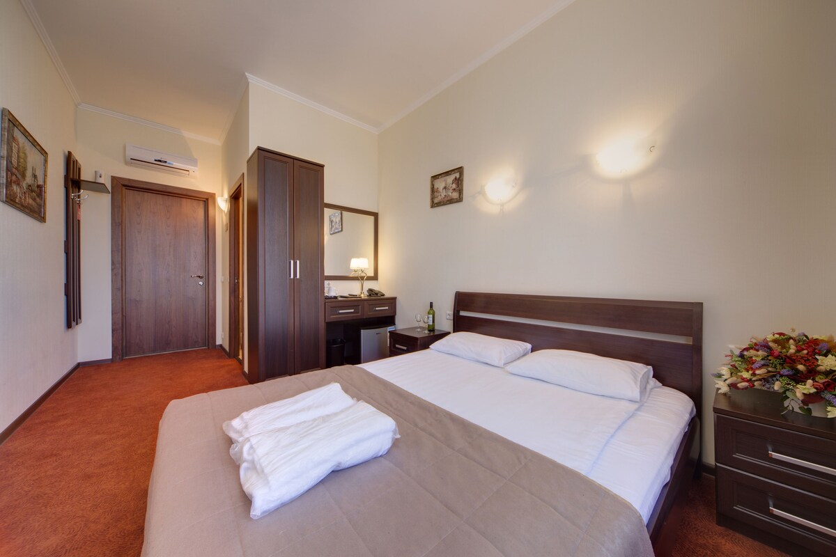 A hotel room with a large bed, a bedside table, a nightstand, a desk with a chair, a mirror, and a wall-mounted air conditioner.