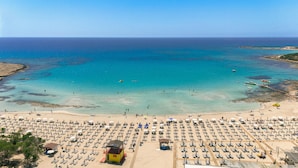 A beach with rows of sunbeds and umbrellas, a lifeguard tower, and boats in the water. - Olympic Lagoon Resort – Ayia Napa (Ayia Napa)