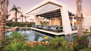 A modern outdoor dining area with a pool, surrounded by palm trees and greenery. - Olympic Lagoon Resort – Ayia Napa (Ayia Napa)