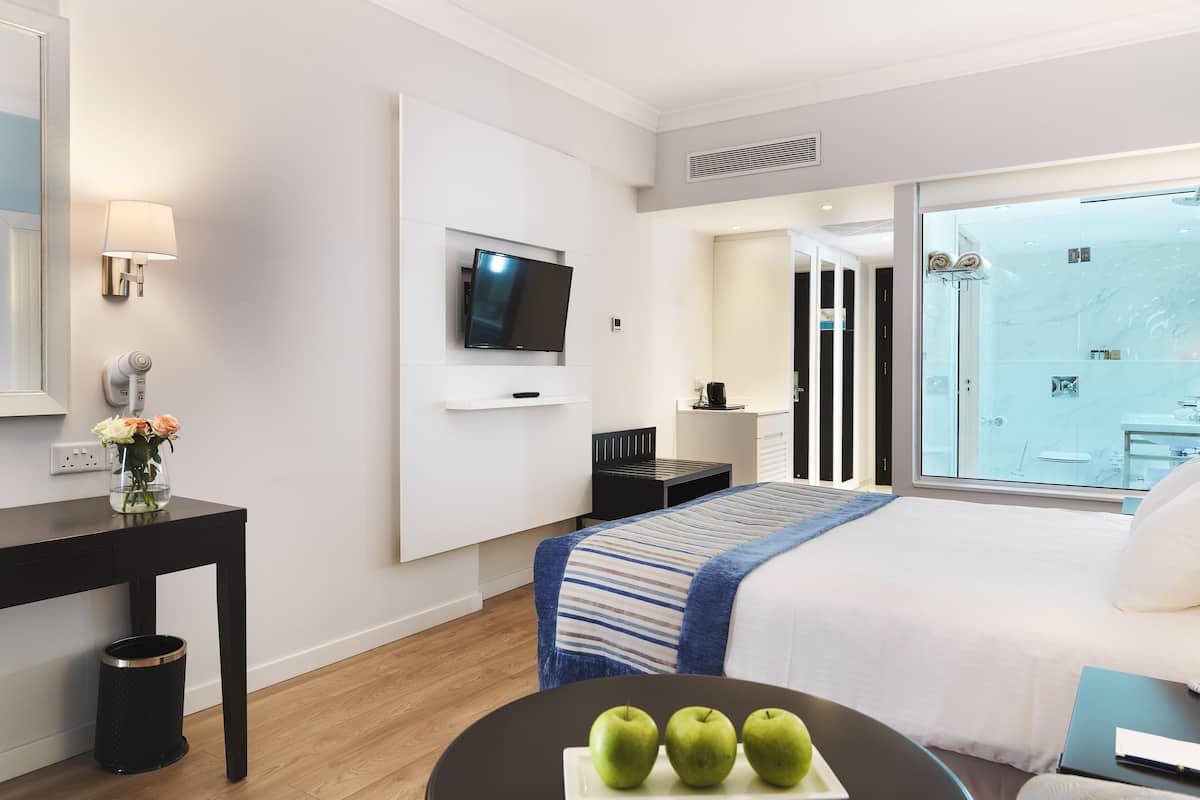 Moderni hotellihuone, jossa on suuri sänky, taulutelevisio, pieni pöytä, jossa on omenoita, ja ikkunasta avautuva näkymä kaupunkiin.