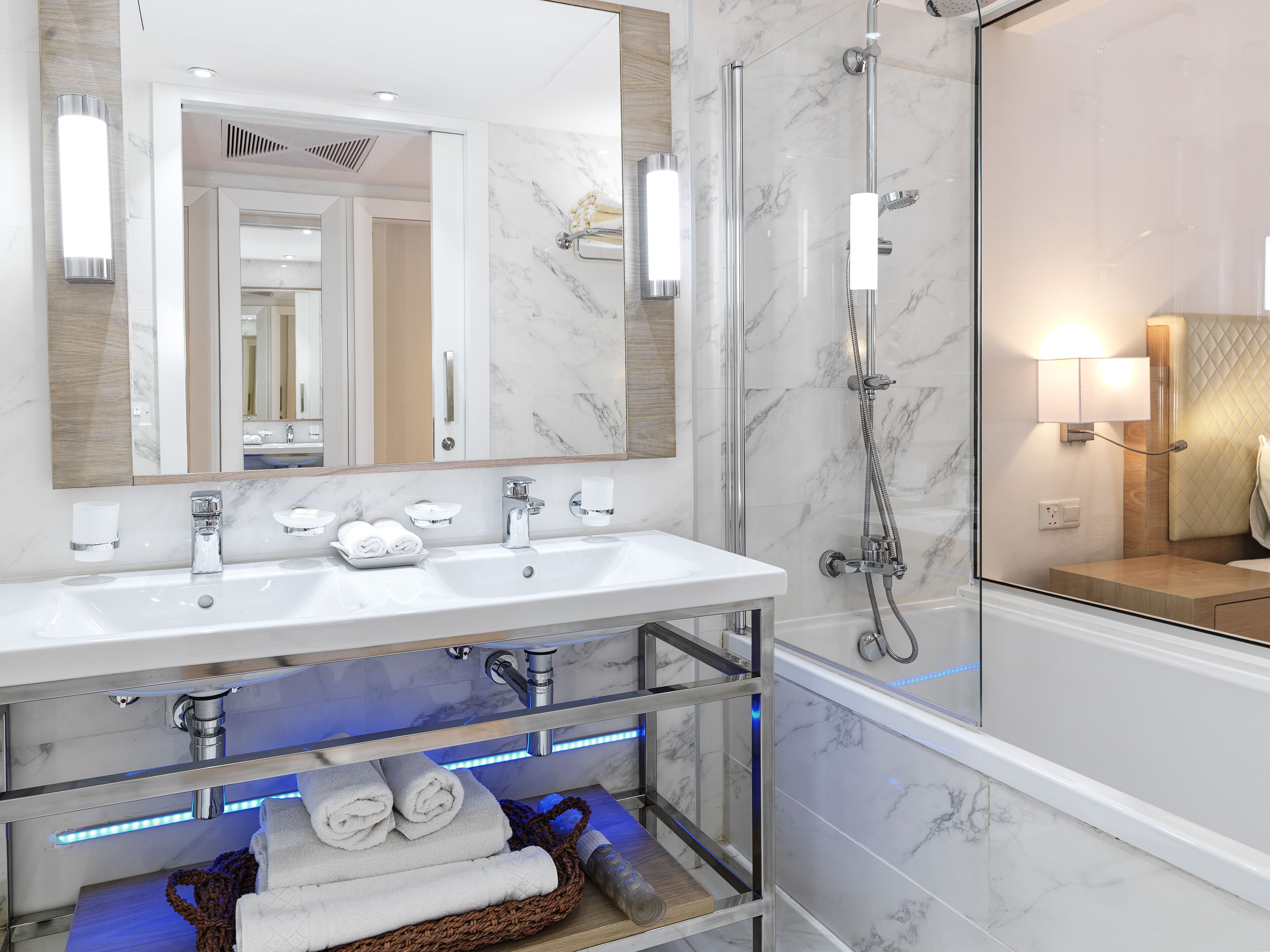 A modern bathroom with a large mirror, two sinks, a glass shelf with towels, and a shower area.