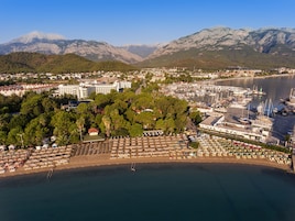 Vista aérea de un pueblo costero con un puerto deportivo, playa con sombrillas y montañas al fondo.