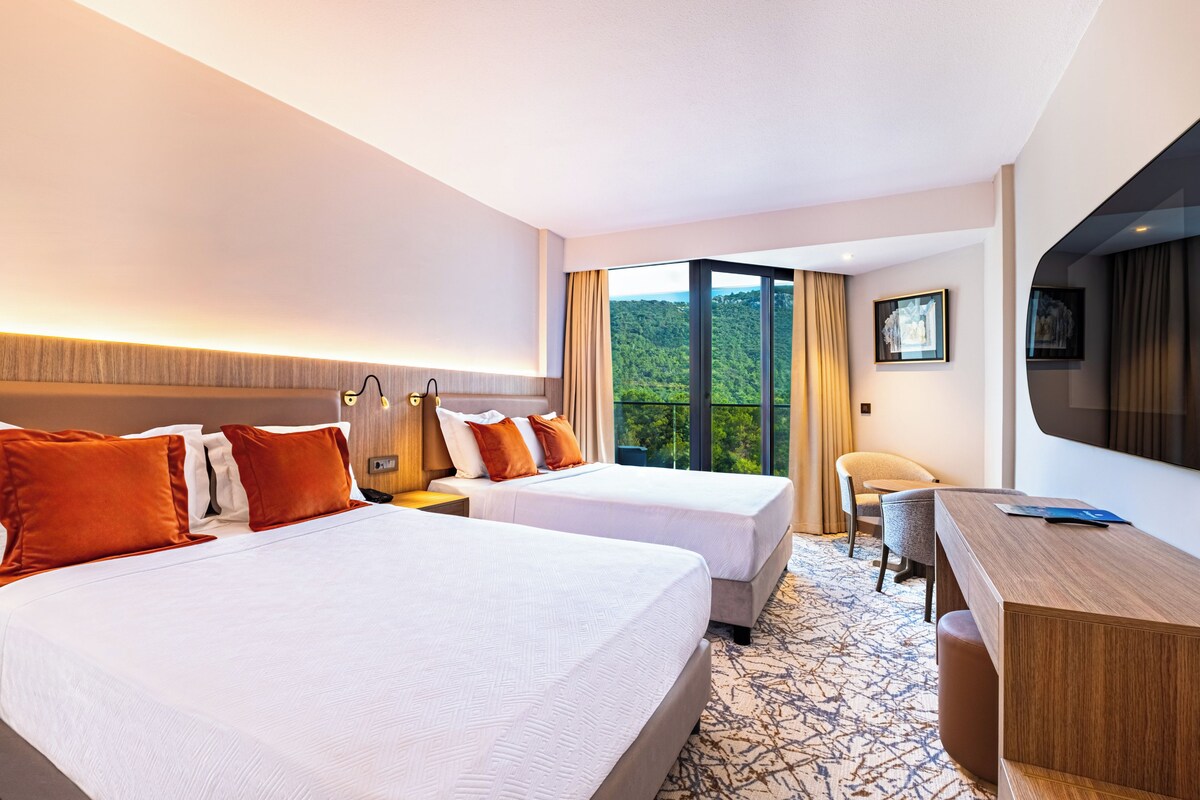 A hotel room with two beds, a desk, a chair, and a large window offering a view of greenery.