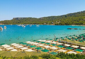 Uma praia com vários guarda-sóis e espreguiçadeiras, mar azul cristalino e barcos ao longe.