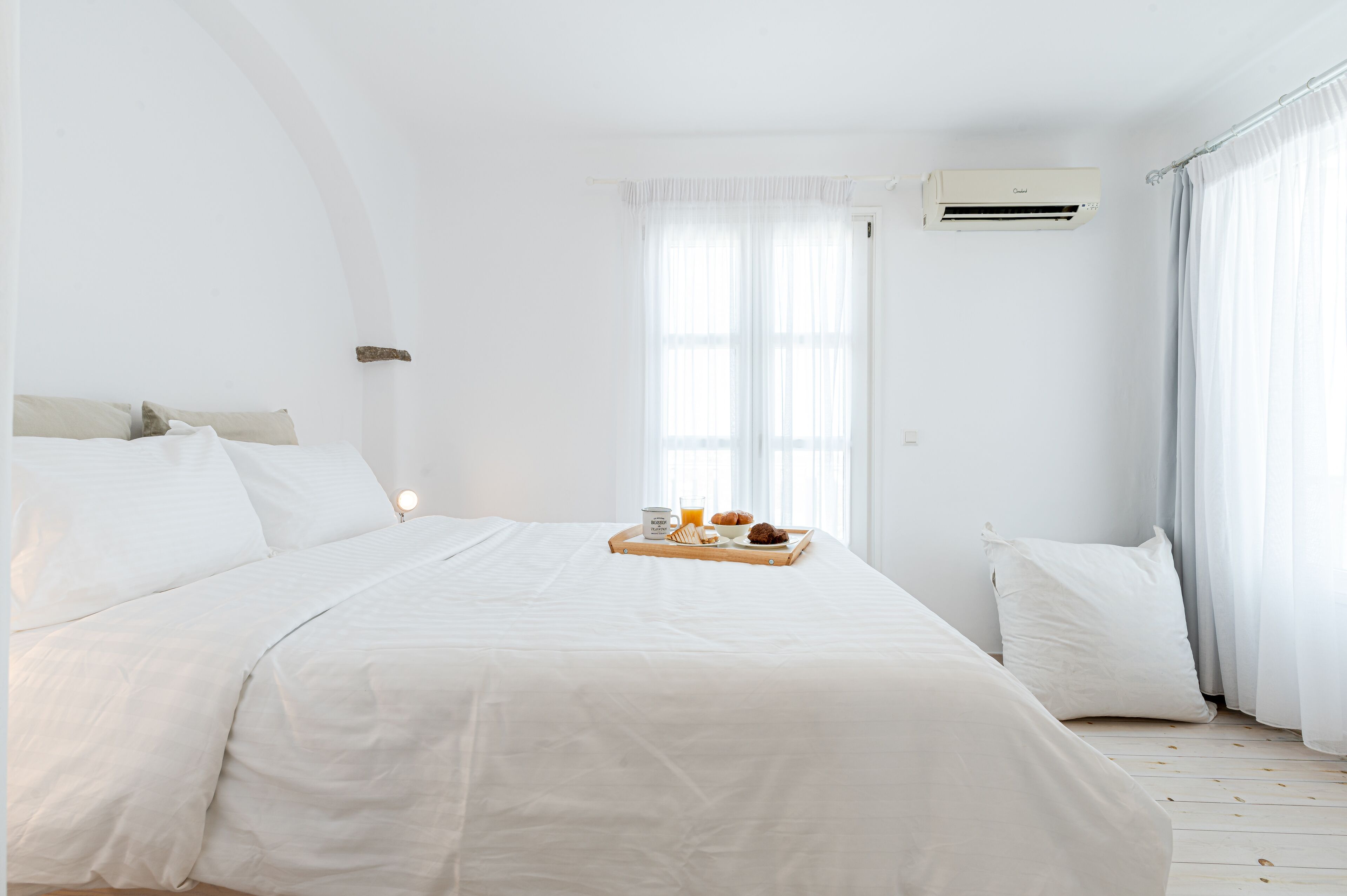 A white bedroom with a bed, a tray with breakfast items, and an air conditioning unit.