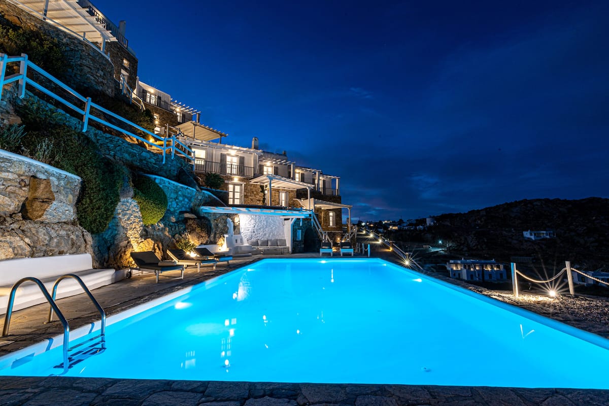 A swimming pool with a stone deck, a stone wall, and a building with multiple balconies and lights.