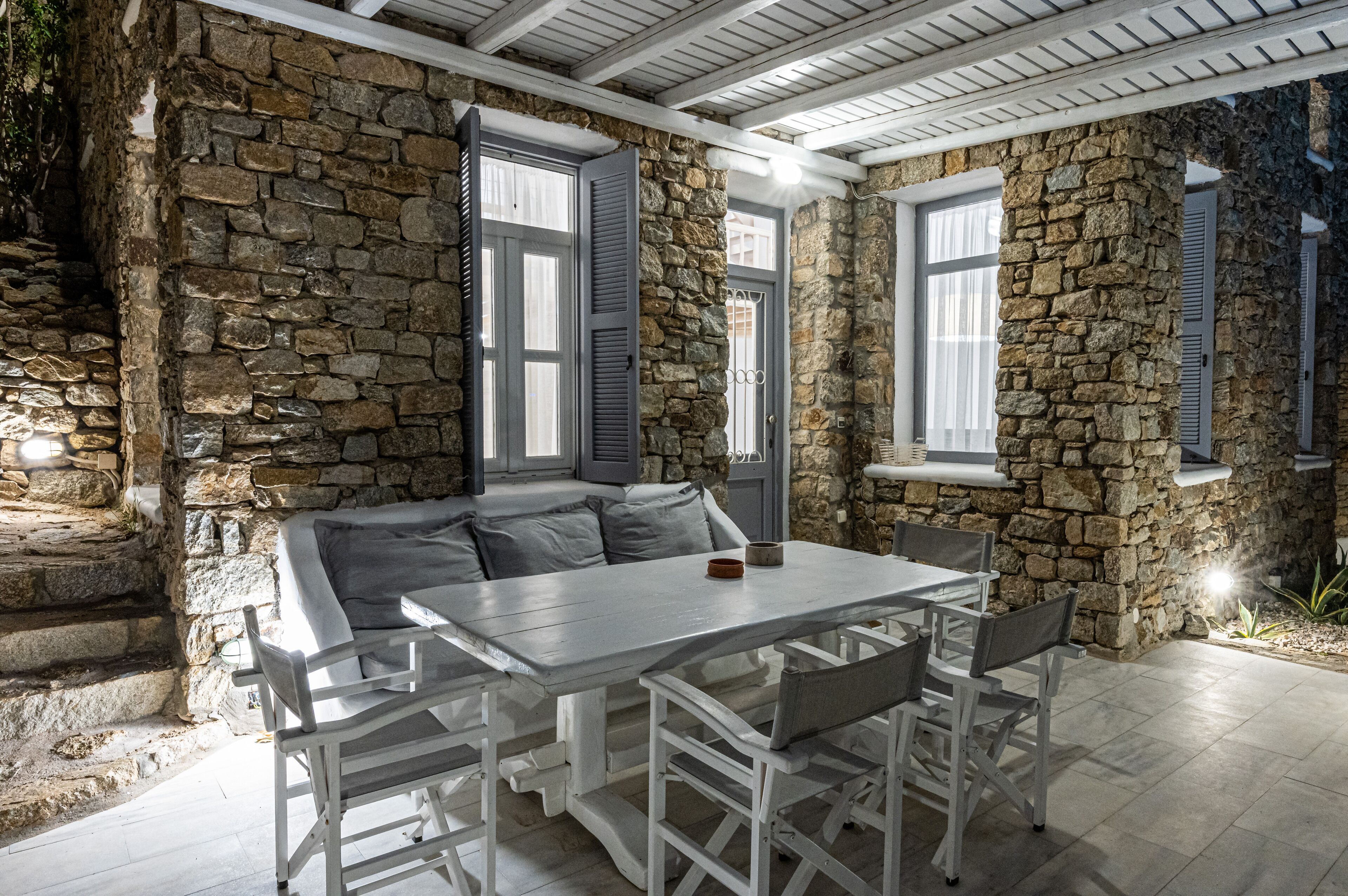 A stone-walled room with a white table and chairs, a bench, and a door with shutters.