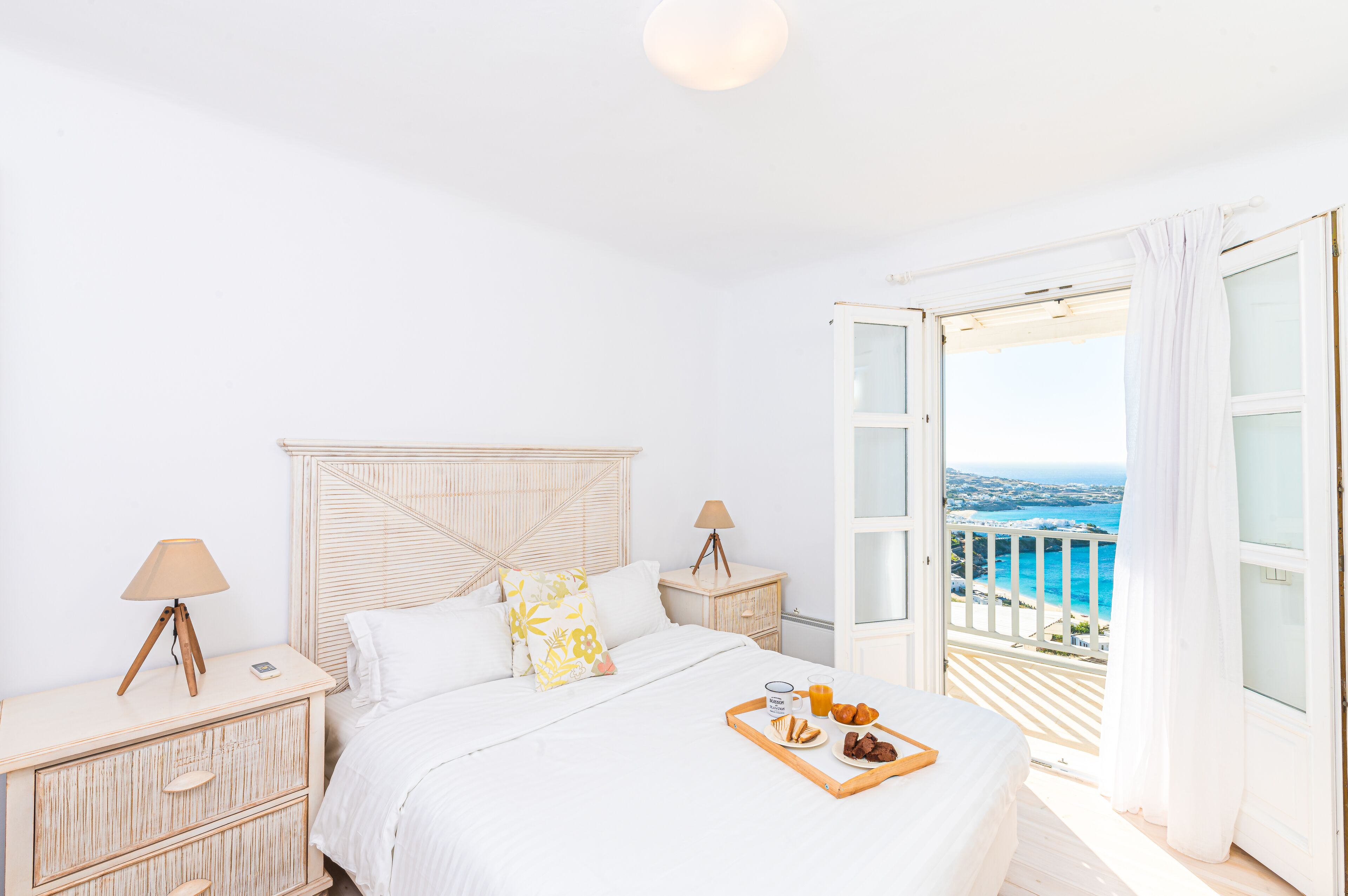 Une chambre à coucher avec un lit, des tables de chevet, un plateau avec de la nourriture, et une vue sur la mer par une porte ouverte.
