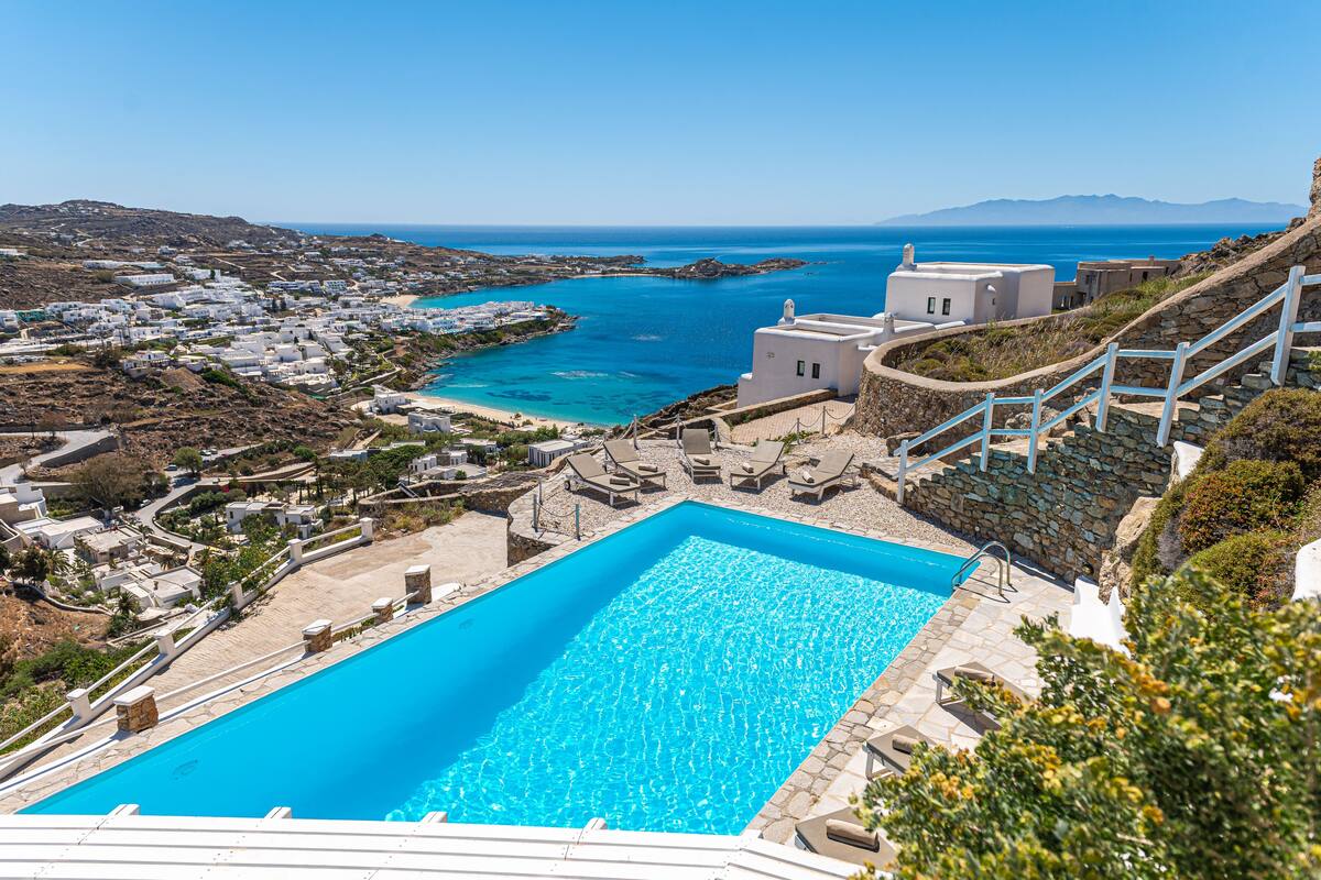 A pool area with a clear blue pool, stone steps, and a view of the sea and buildings in the background.