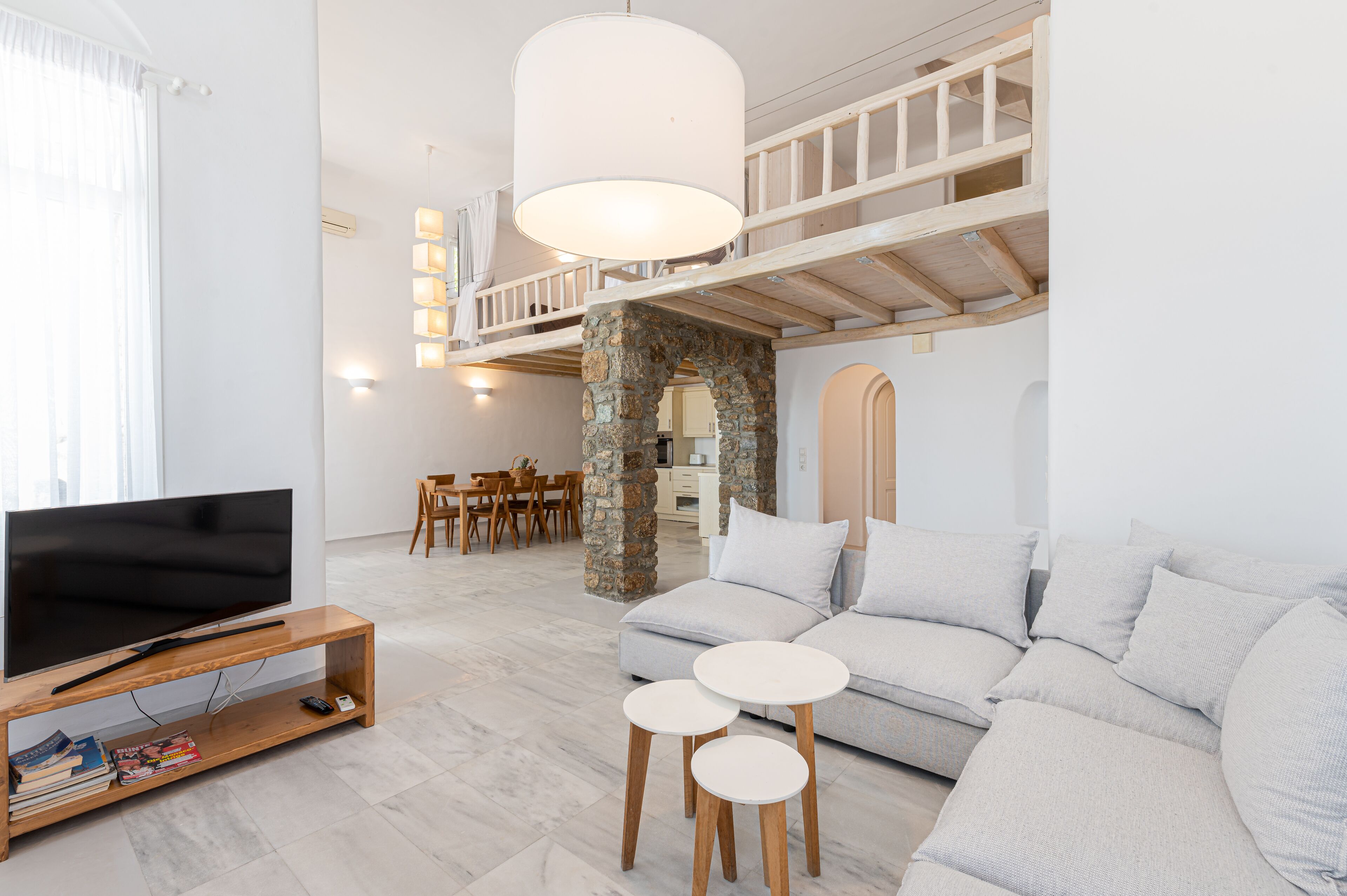 Una sala moderna con televisión, sofá y dos mesitas. Se ve una escalera que sube a un nivel superior con balcón.