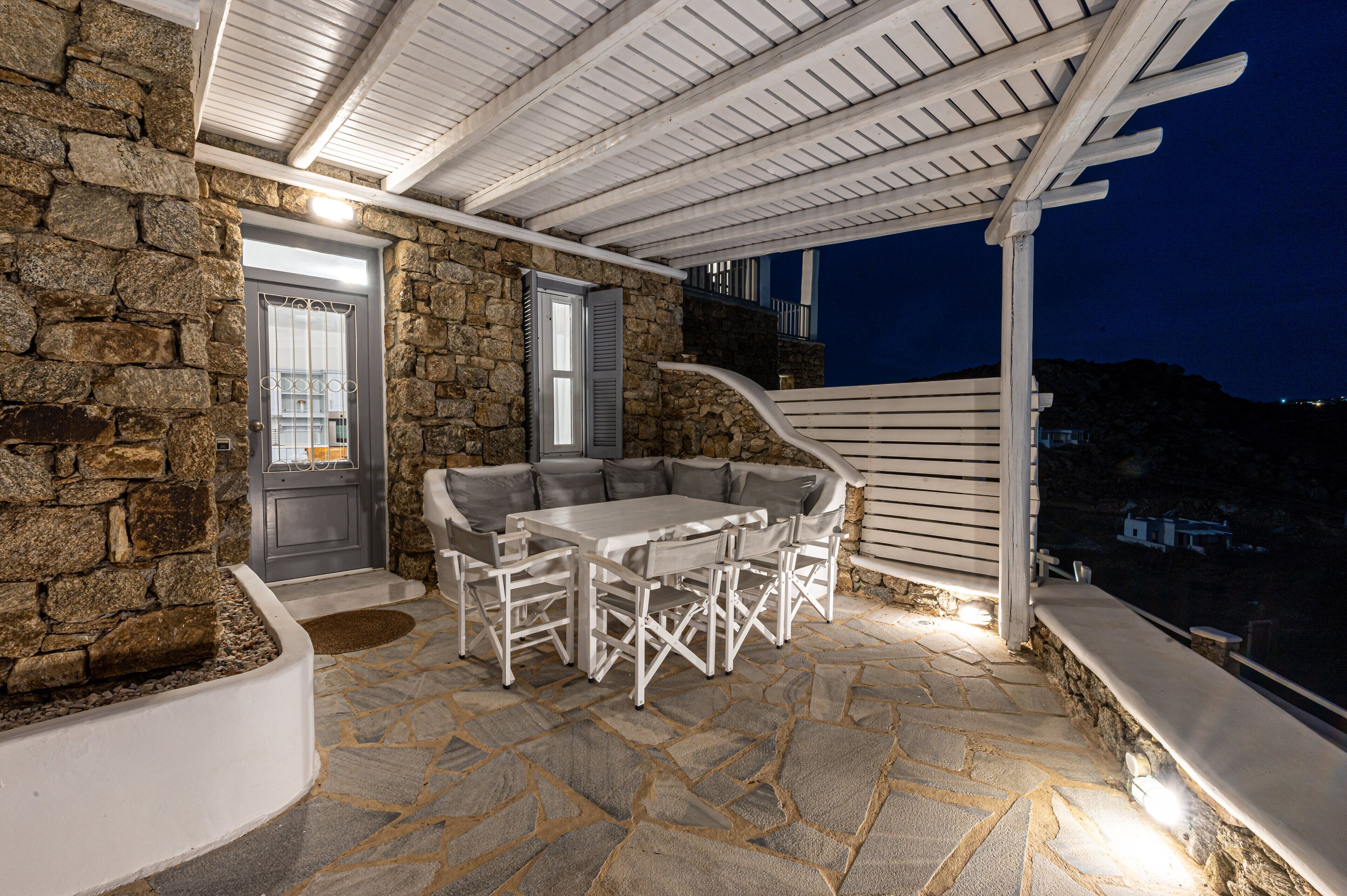 A stone patio with a white table and chairs, a door with a window, and a white pergola.