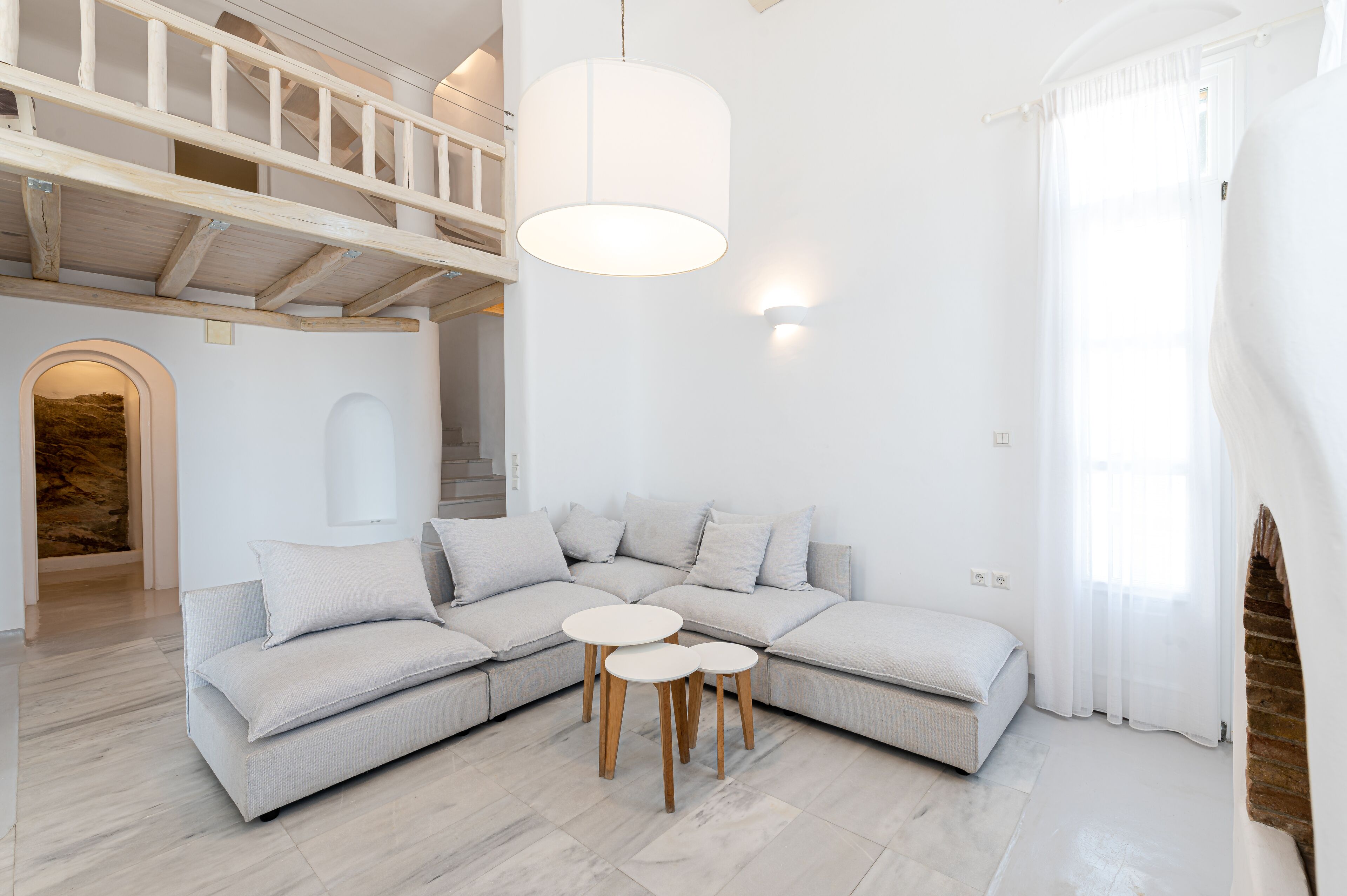 Een moderne woonkamer met een hoekbank, twee ronde houten tafels, een hanglamp en een trap naar een verdieping erboven.