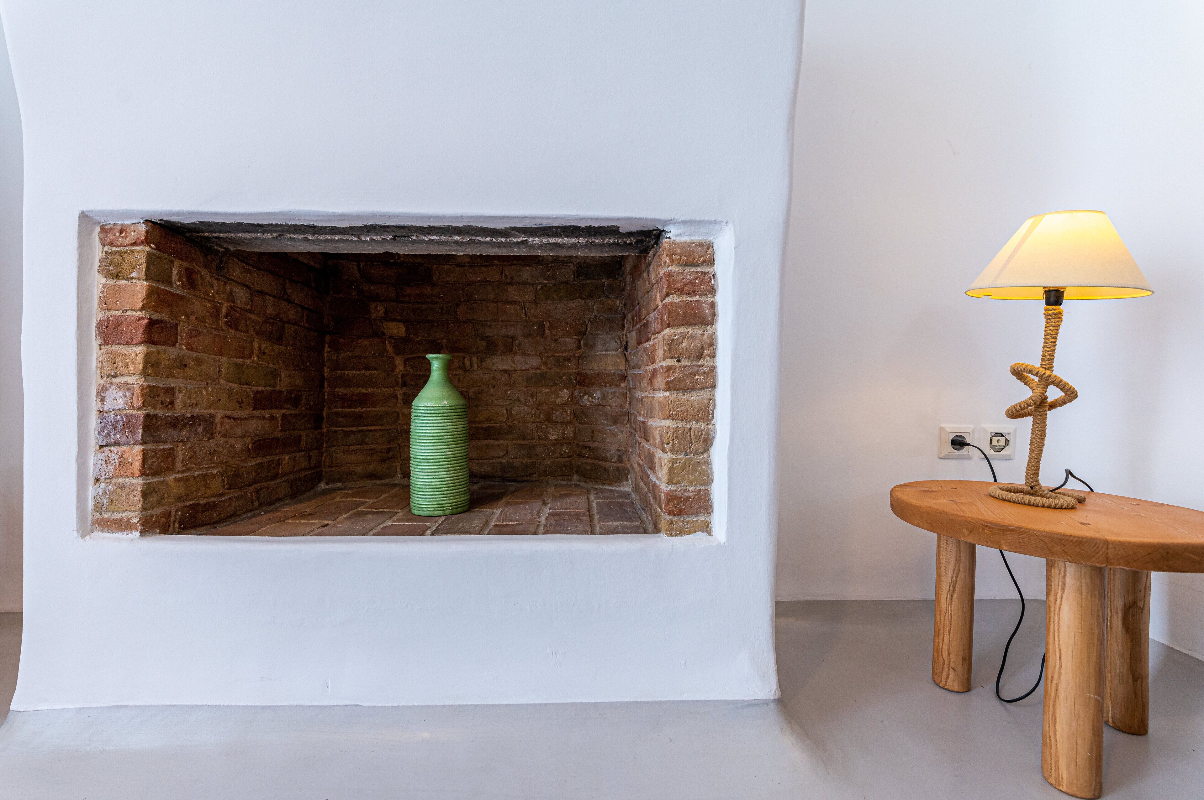 Una chimenea, un jarrón verde, una mesa de madera con una lámpara y una pared blanca.