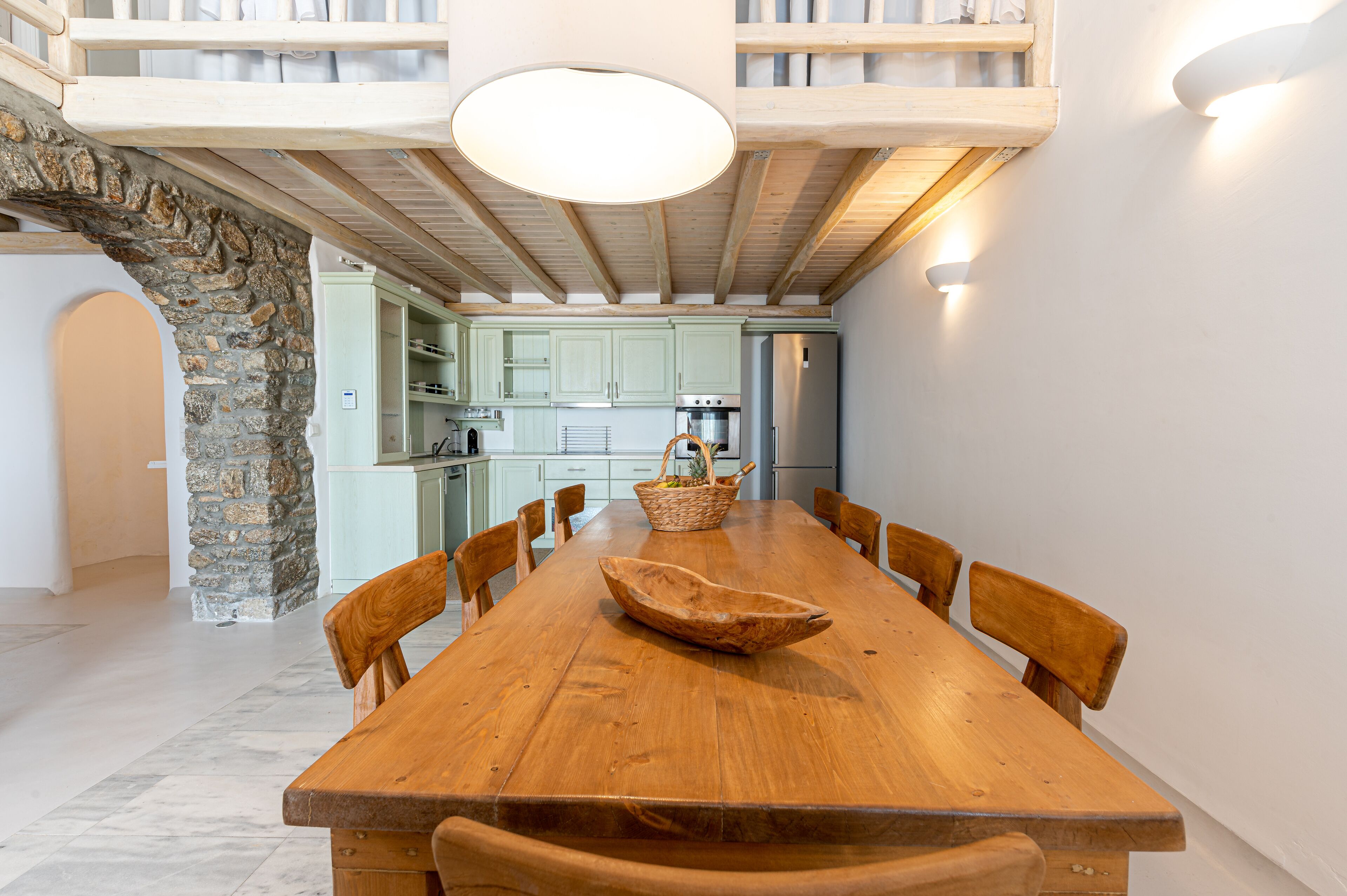 A spacious dining area with a long wooden table, surrounded by chairs, and a stone wall on one side.