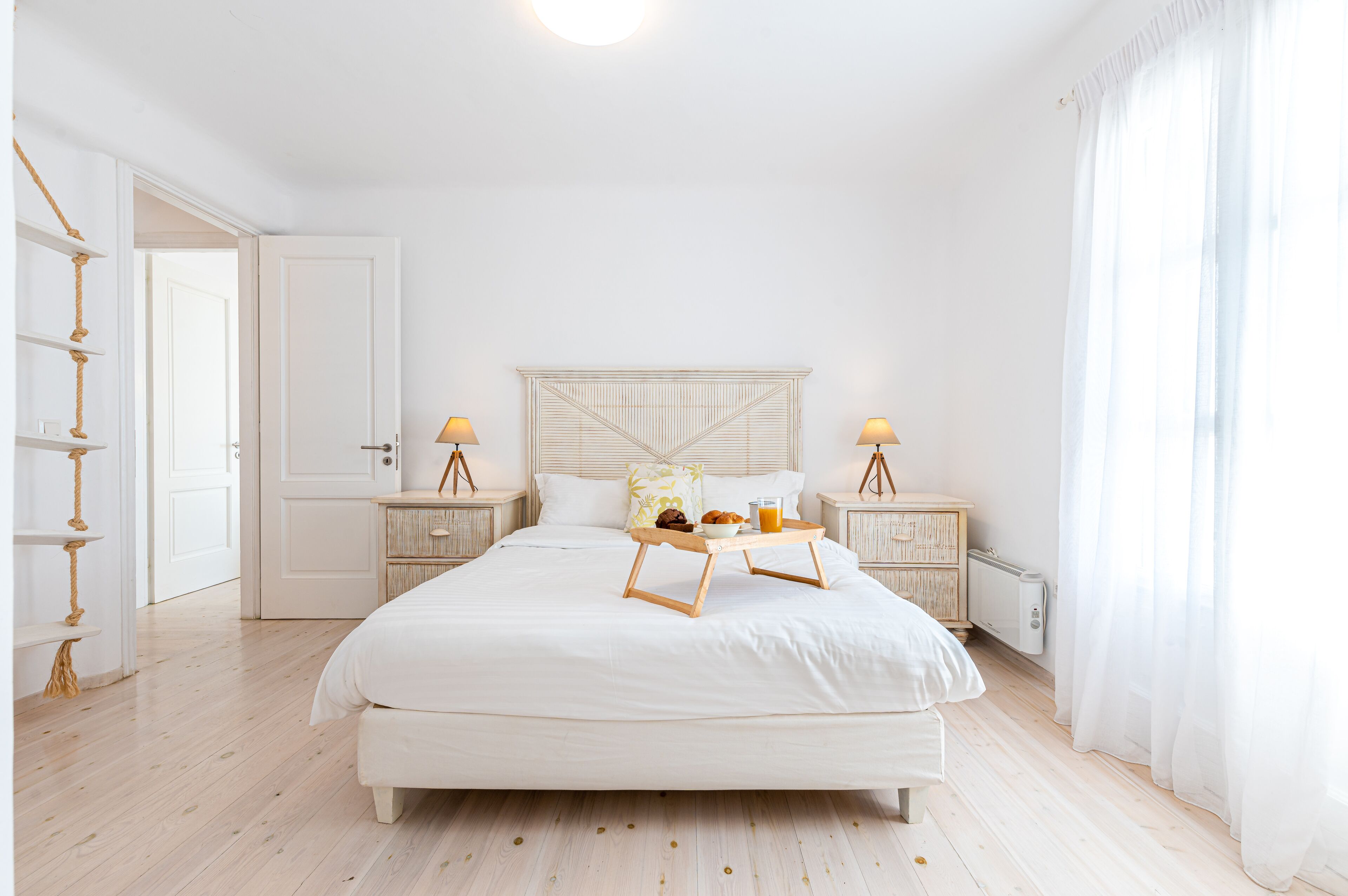 A spacious bedroom with a large bed, bedside tables with lamps, a bedside table with a tray of breakfast, and a view of the outside through sheer curtains.