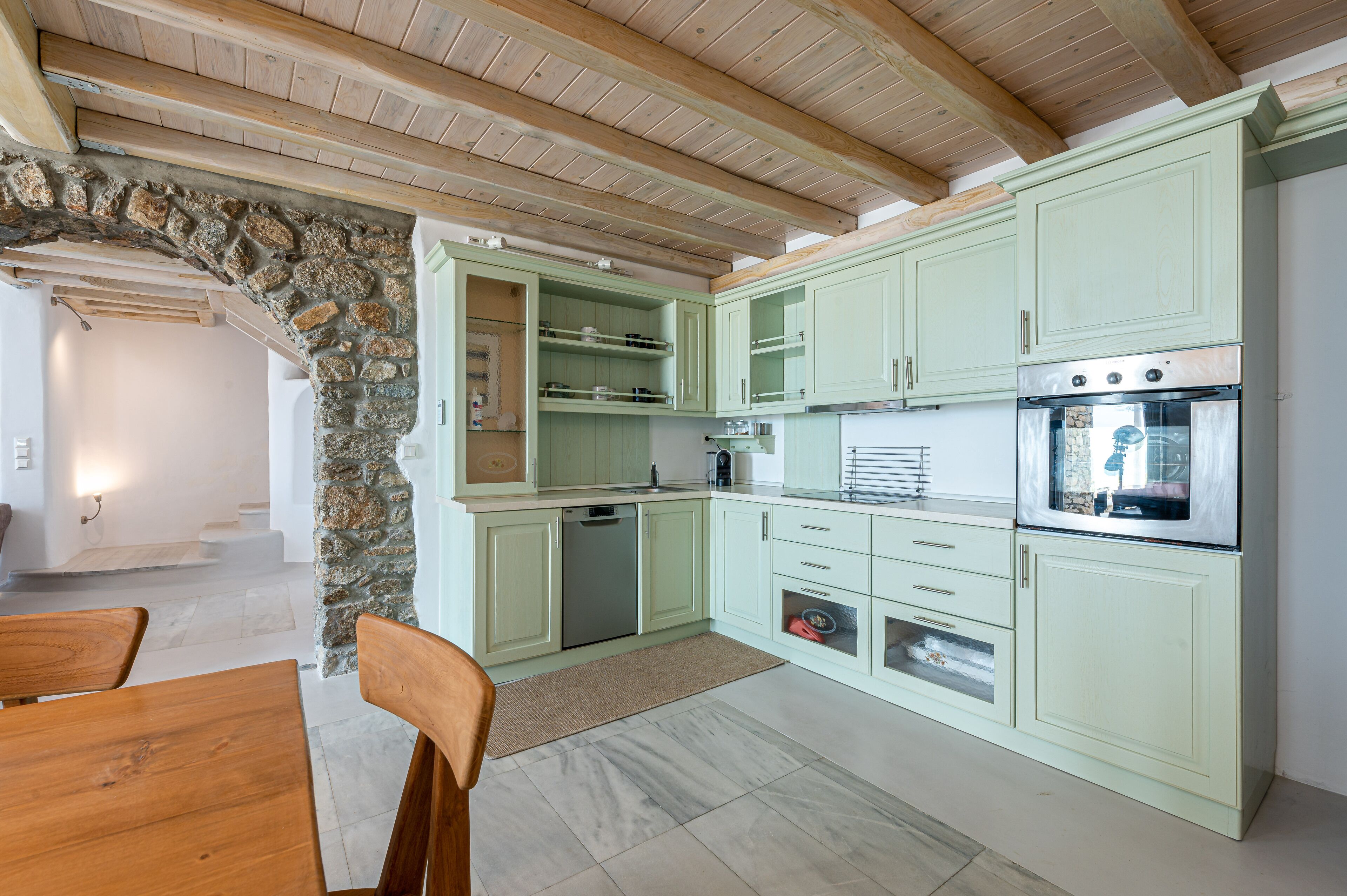A kitchen with light green cabinets, a stone archway, wooden ceiling beams, and a built-in oven.