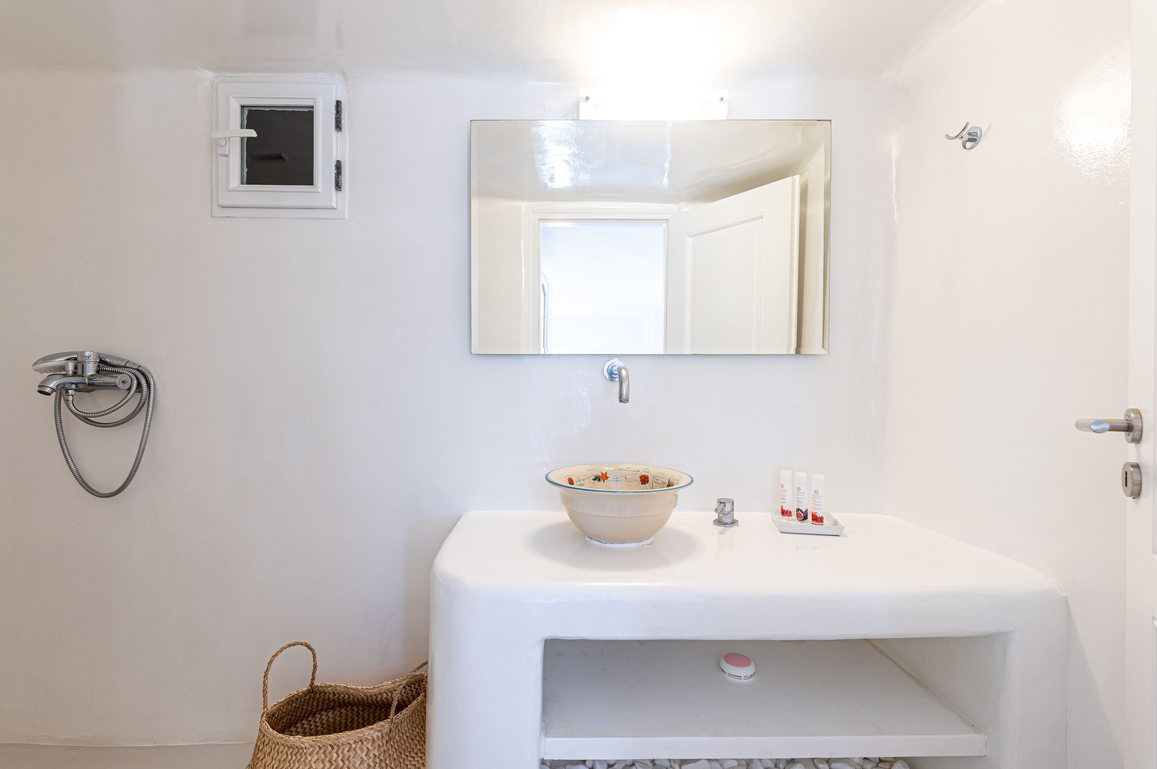Une salle de bain avec un lavabo blanc, un miroir, une pomme de douche et une petite fenêtre.