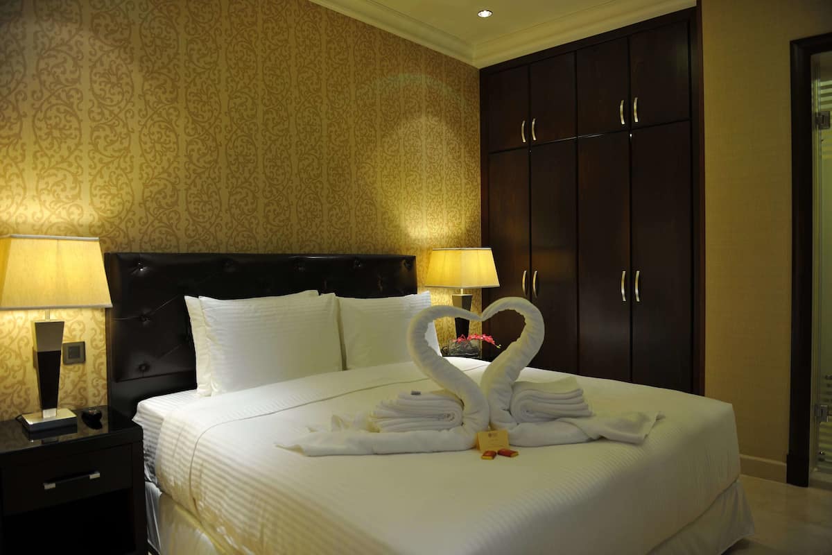 A hotel room with a neatly made bed, two bedside lamps, a dark wooden wardrobe, and a decorative wall pattern.