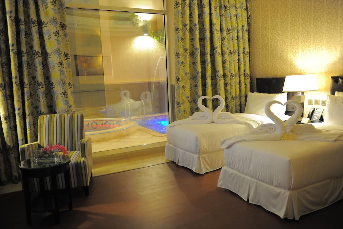 A hotel room with two beds, a glass-enclosed jacuzzi, a chair, and a table with flowers.