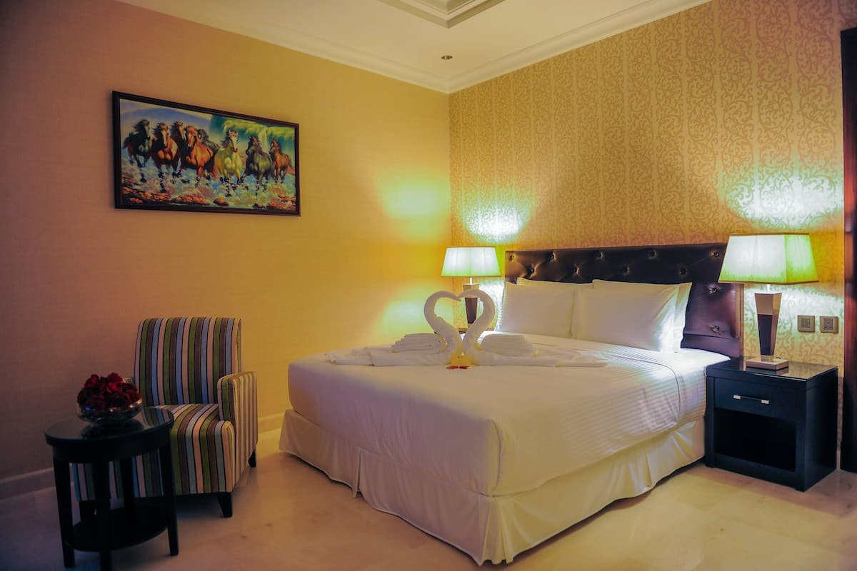 A hotel room with a large bed, two bedside lamps, a striped armchair, a painting on the wall, and a small table with a bowl of flowers.