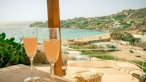 Two glasses of a clear beverage on a table with a view of a beach and hillside buildings. - Super Rockies (Mykonos)