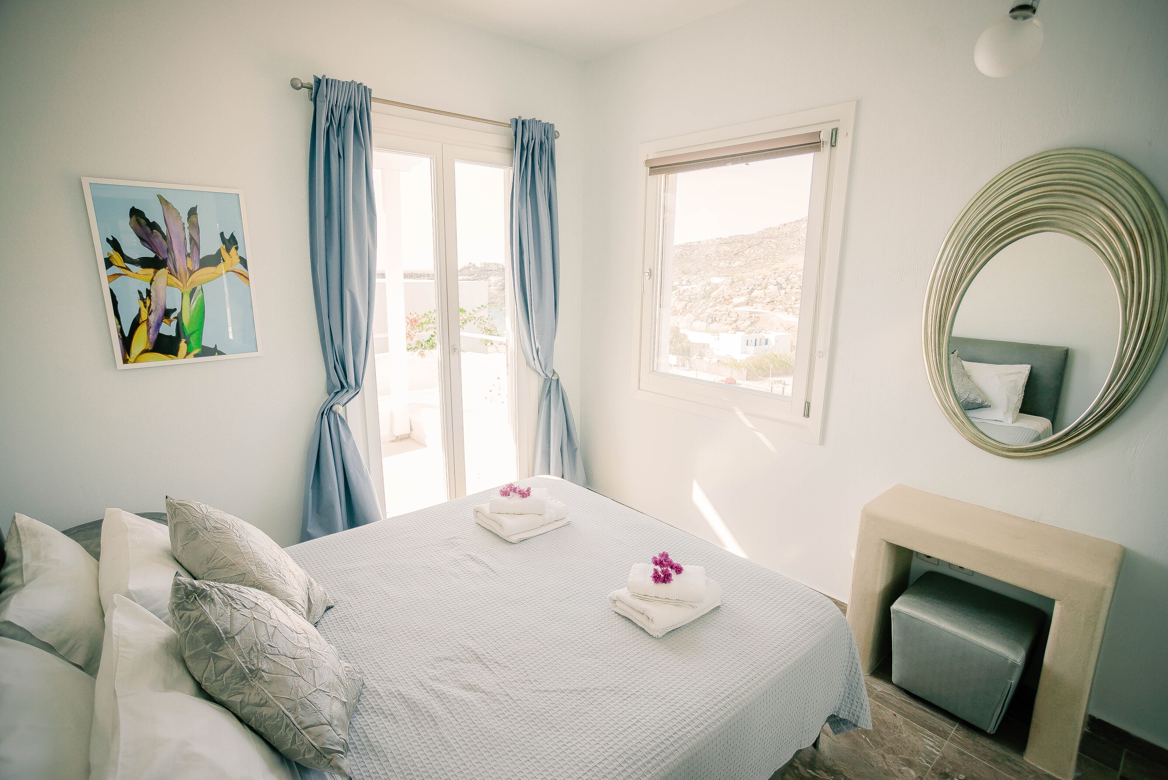 A bedroom with a bed, pillows, a painting, a window with curtains, a mirror, and a fireplace.