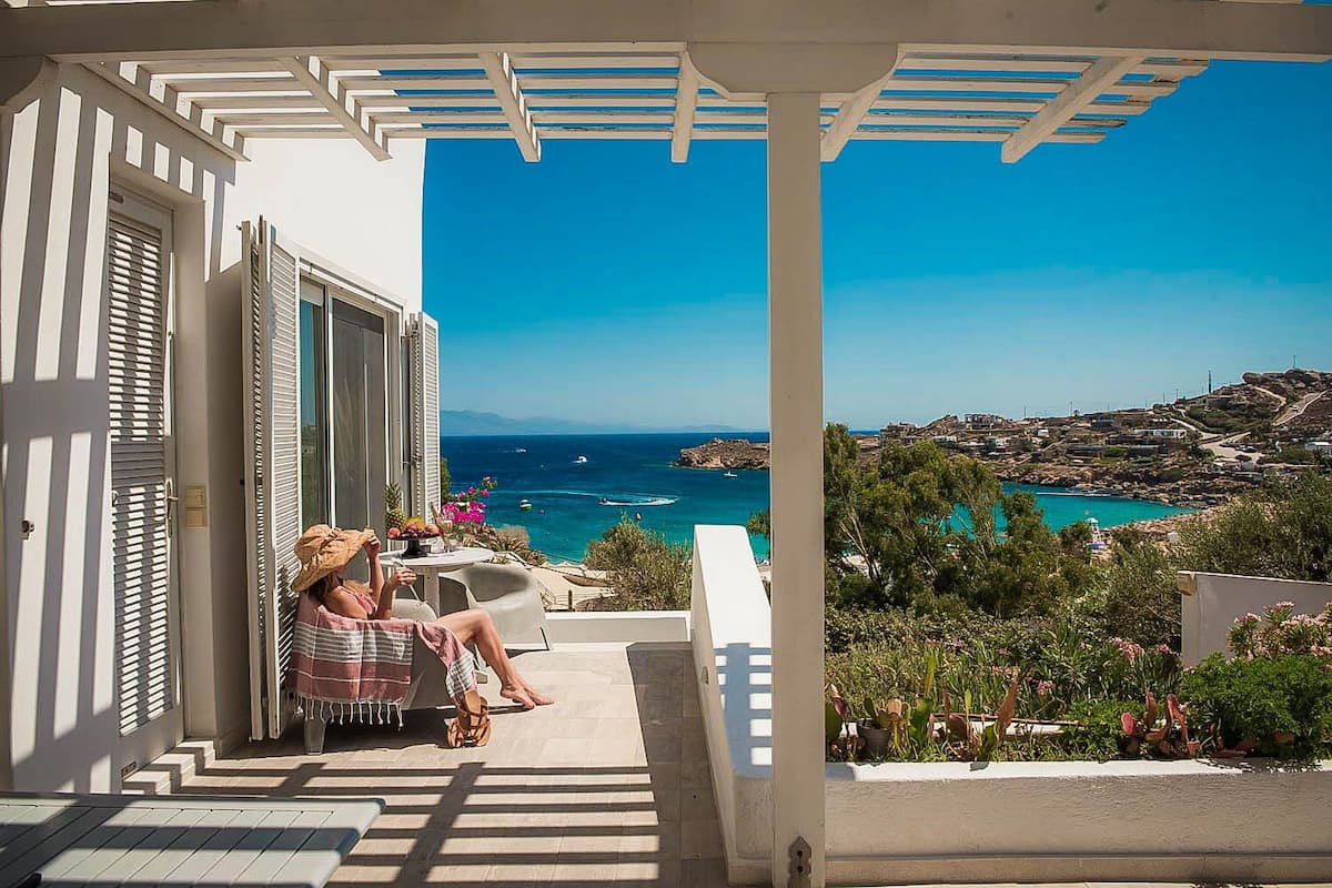 Un balcón con vistas al mar, una persona sentada en una silla y una mesa con una bebida.