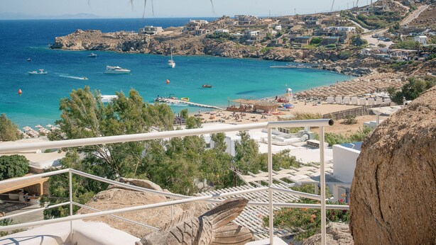 A view of a coastal town with a rocky shoreline, boats, and a clear blue sky. - Super Rockies (Mykonos)