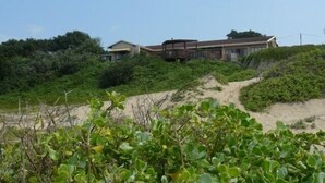 Una propiedad frente al mar con una casa grande y un edificio más pequeño, rodeada de vegetación densa y un sendero de arena que conduce a la playa.