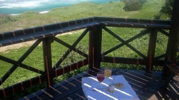 Una terraza de madera con una mesa dispuesta para dos, con vistas a un paisaje costero con olas y vegetación.