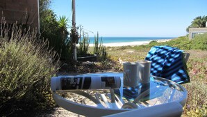 Ein Glastisch mit einem blau-weiß gestreiften Handtuch, zwei Tassen und einer Zeitschrift. Im Hintergrund ist ein Strand und ein Meer zu sehen.