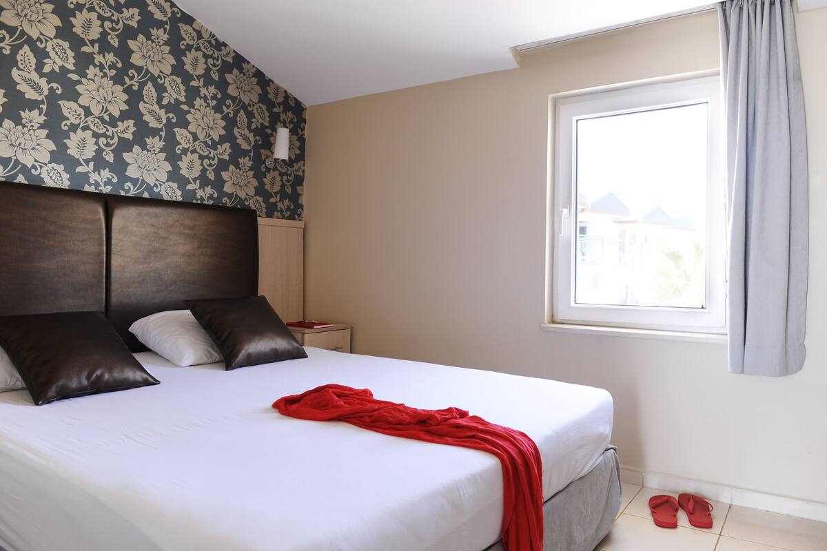 Ein ordentlich bezogenes Bett mit einer weißen und roten Decke, ein Blumenmuster-Kopfstück und ein Paar rote Hausschuhe auf dem Boden.
