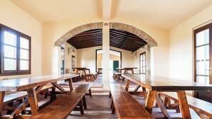 Una lunga sala da pranzo con tavoli e panche in legno, soffitto a volta e ampie finestre.