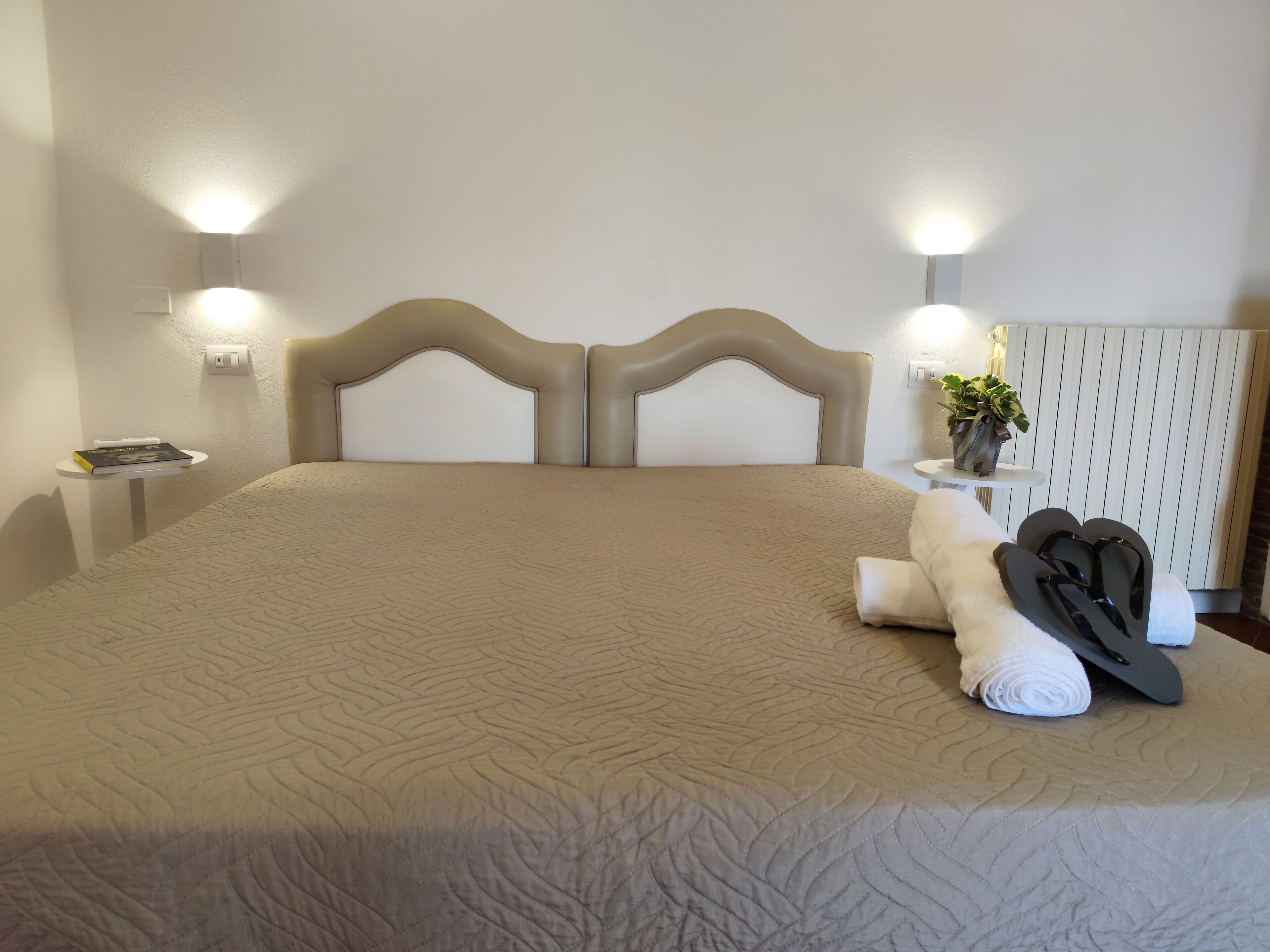 A neatly made bed with a beige quilt, a bedside table with a book, and a wall-mounted lamp.