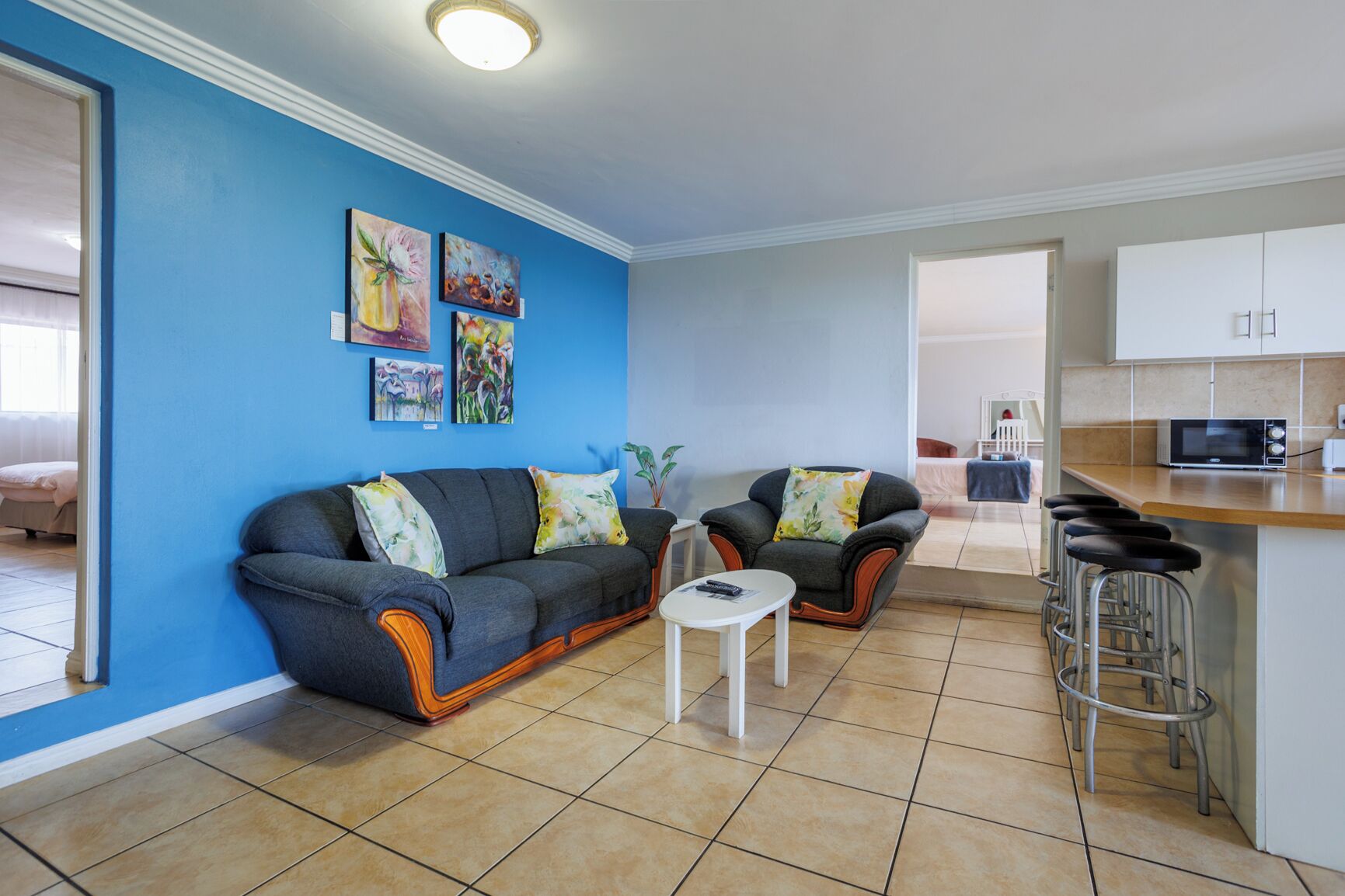 Een woonkamer met een bank, fauteuils, een kleine tafel, en een keukenhoek met een magnetron en barstoelen.