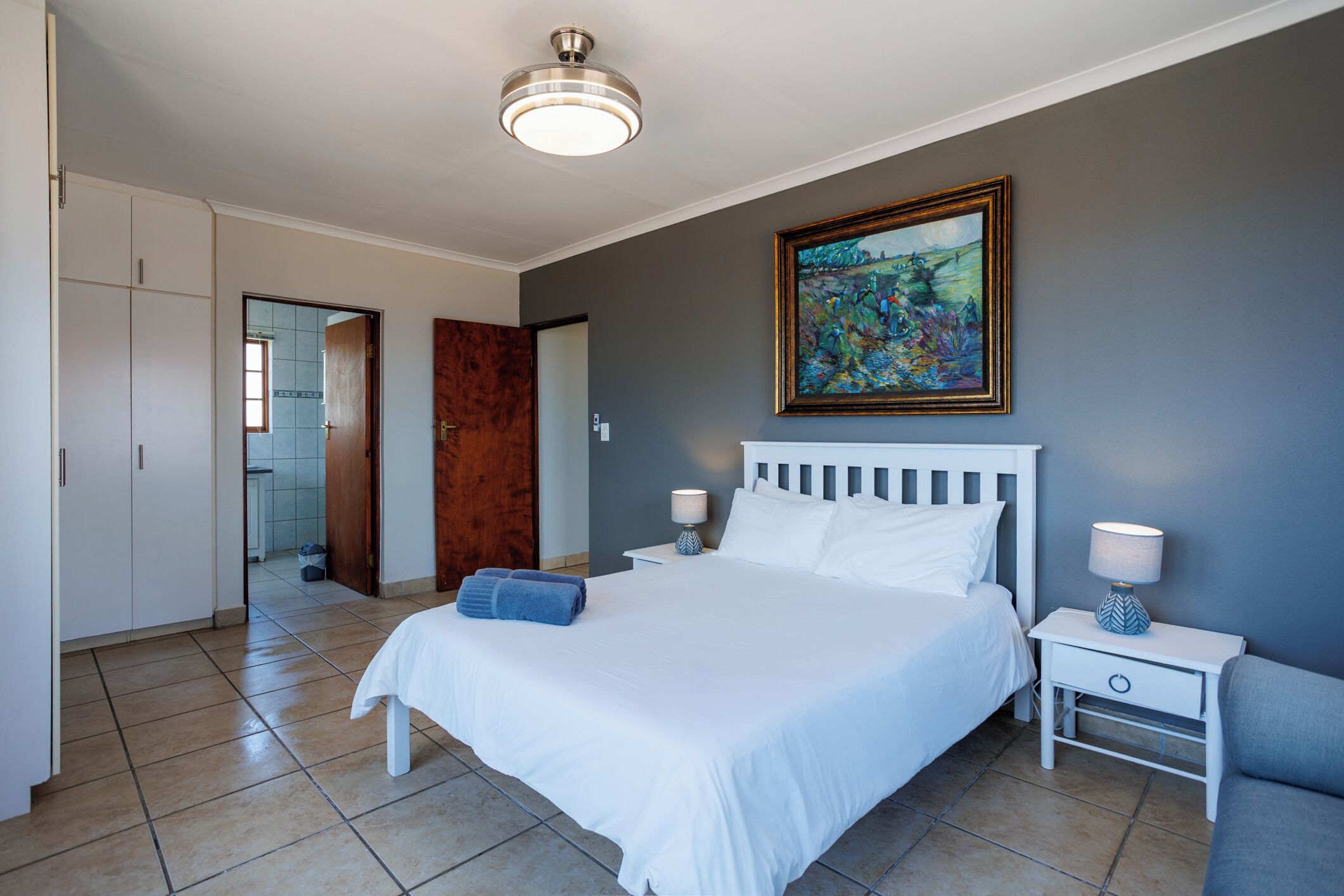 A hotel room with a large bed, two bedside tables, a nightstand, a wardrobe, and a painting on the wall.