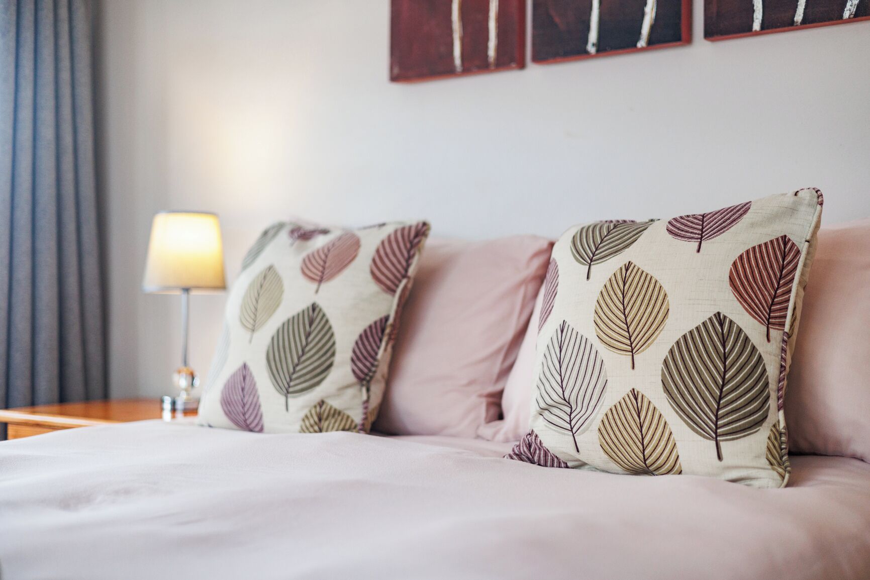 Una cama con dos almohadas de estampado de hojas, una lámpara de noche y una pared con tres cuadros.