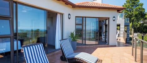 A modern outdoor patio with two striped lounge chairs, a glass door leading to a covered area, and a view of the landscape.