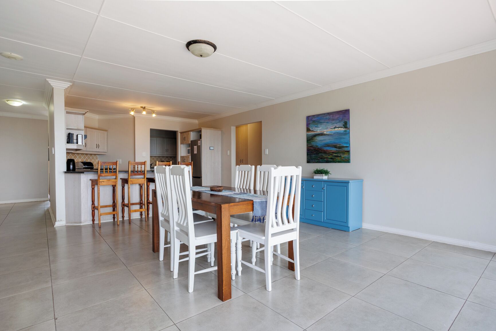 A dining area with a wooden table and white chairs, a kitchen in the background, and a blue cabinet.