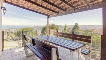 A covered outdoor seating area with a bench and a table, overlooking a cityscape and hills.