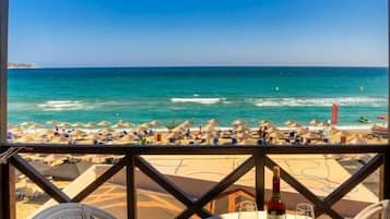 Mesa com garrafa e copos, com vista para uma praia com guarda-sóis e o mar.