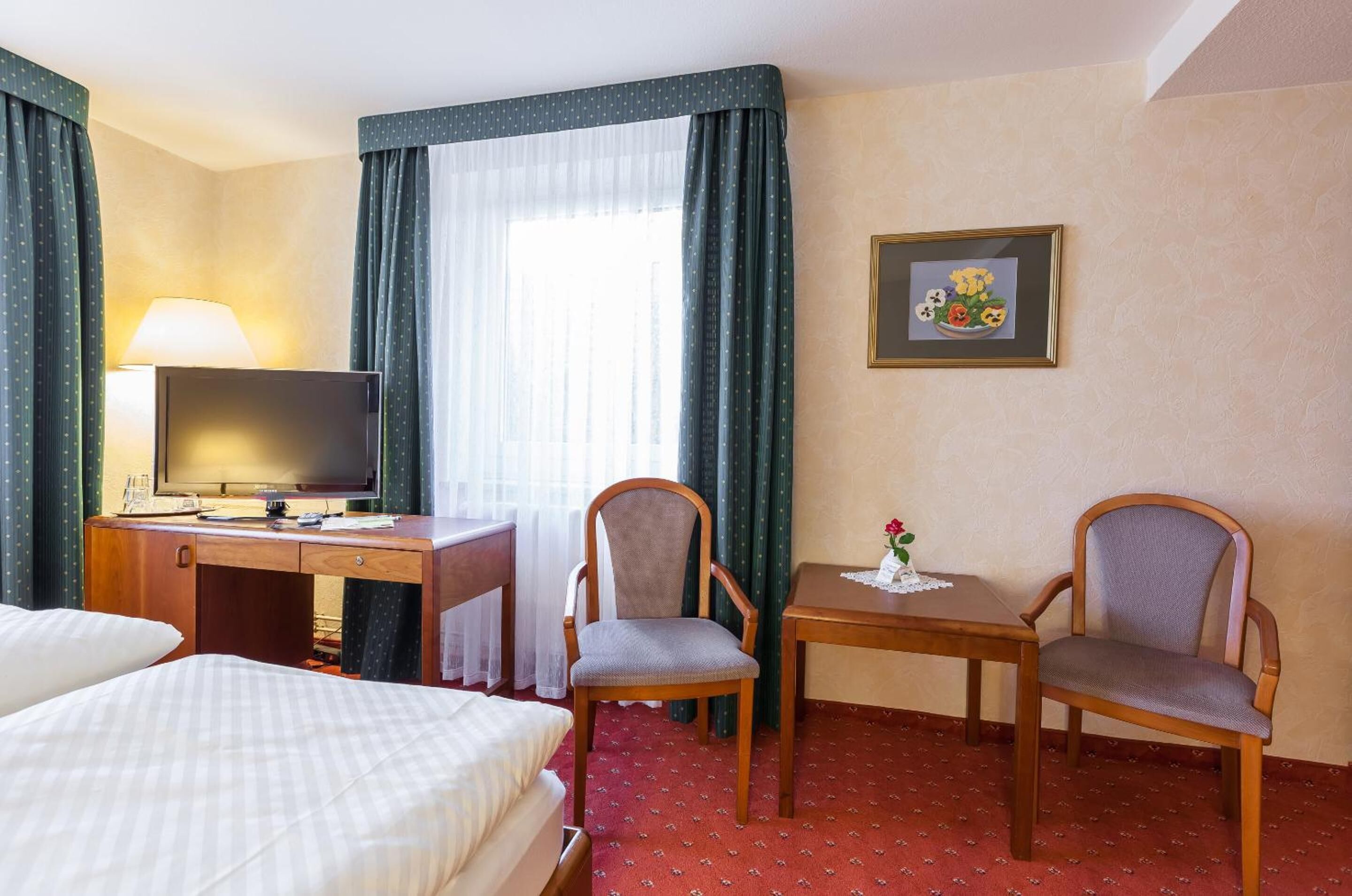 Une chambre d’hôtel avec un lit, un bureau avec une télévision, deux chaises, une petite table avec une fleur et un tableau encadré au mur.