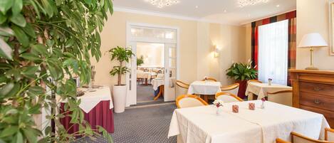 A hotel dining area with tables, chairs, potted plants, and a view into the dining room.