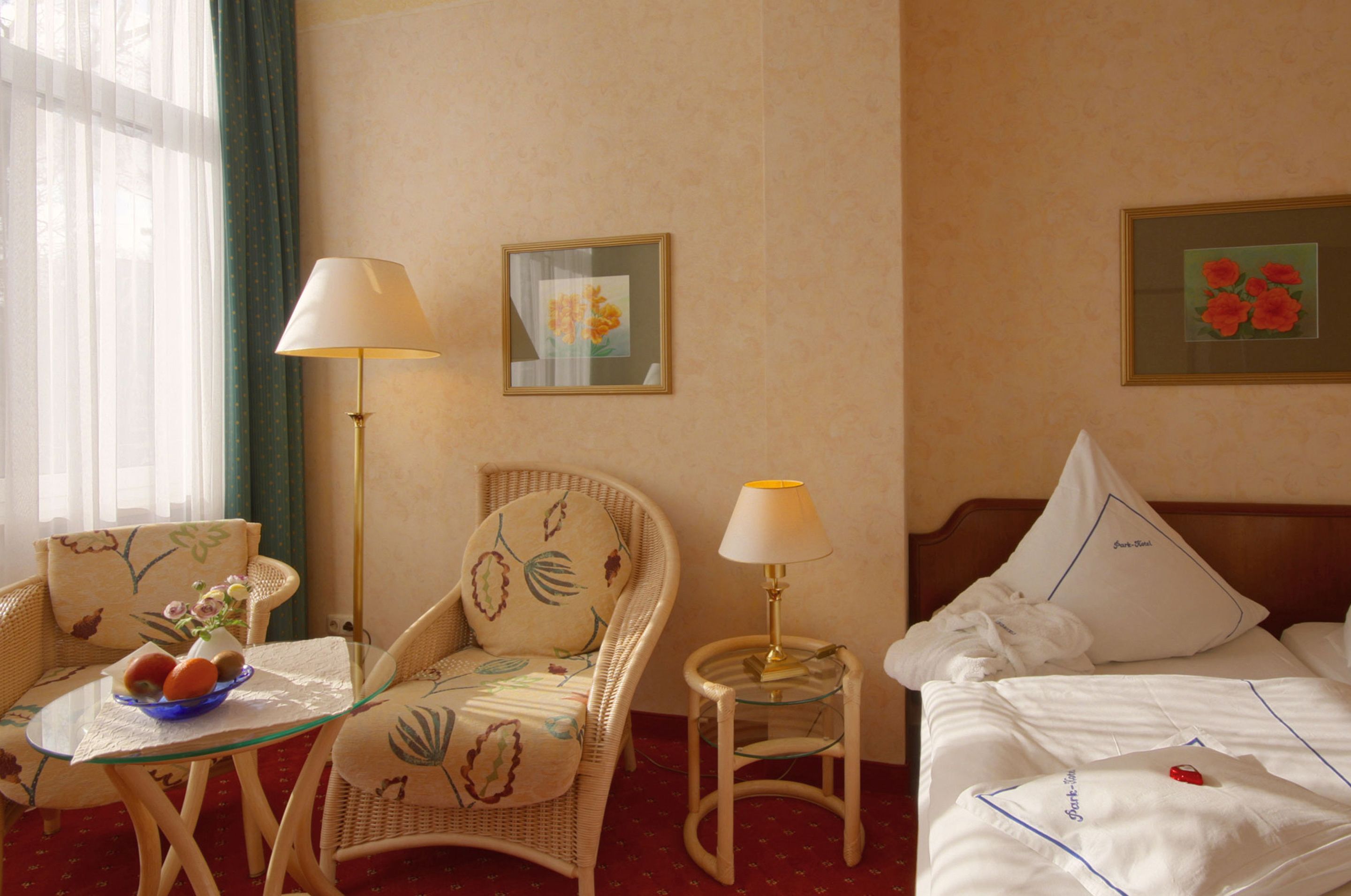 A hotel room with a bed, two chairs, a small table with a bowl of fruit, a lamp, and two framed pictures on the wall.