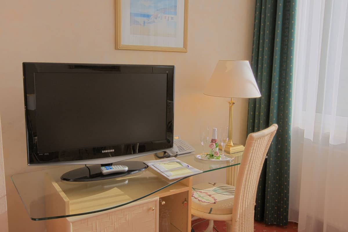 A hotel room with a flat-screen TV, a glass desk, a lamp, a chair, and a window with curtains.
