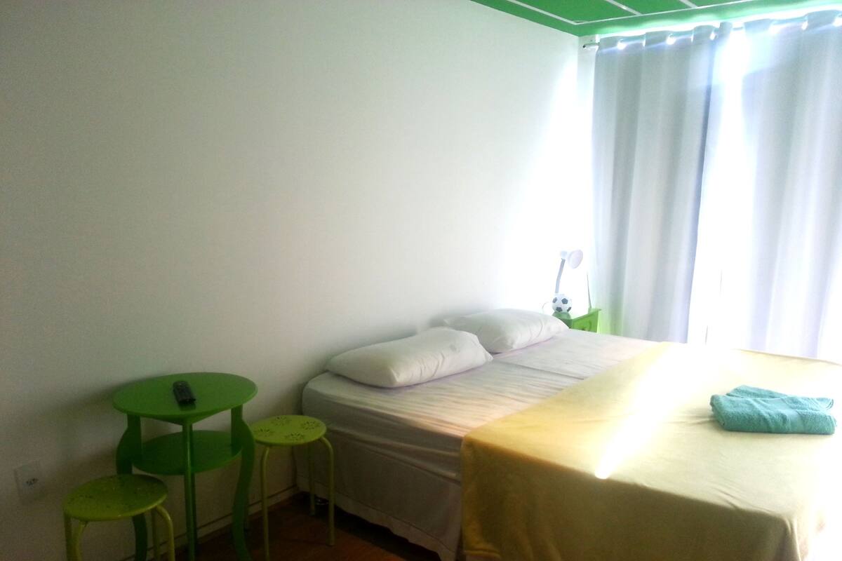 A small, clean room with a bed, a green chair, a small table, and a window with curtains.